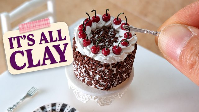 A hand holding a tiny Black Forest cake made from polymer clay on a miniature cake stand, with the words It's all clay