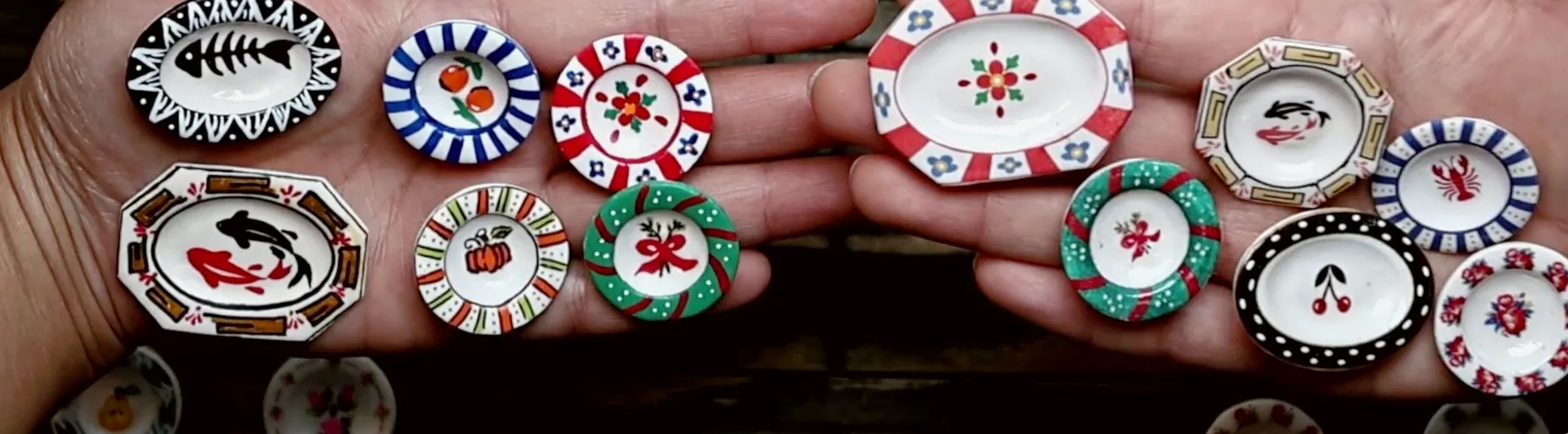 Two open hands holding a dozen small painted plates, including a fishbone oval, koi, oranges, a pumpkin, a bow, a lobster and black-and-gold cherries