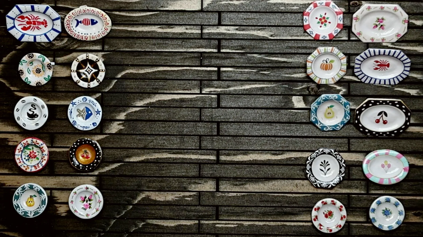 Two columns of small painted miniature plates laid out on a dark wooden board: ovals, octagons and rounds with lobsters, cherries, a pumpkin, a pear, sunflowers and more